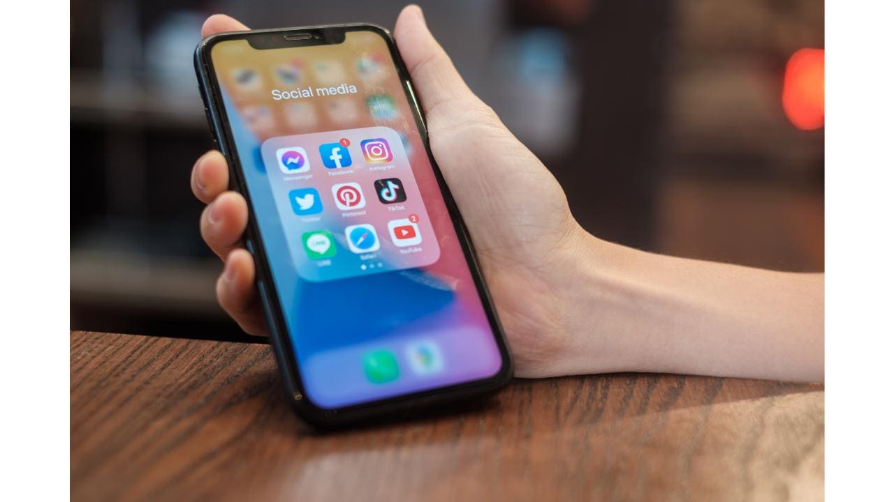 ソーシャルメディアのアプリアイコンが表示されたスマートフォンを持つ手。木目調の背景。