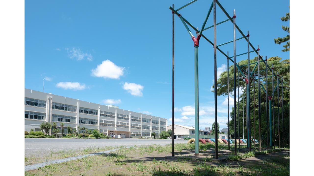 空の下に広がる学校の校庭と校舎、手前に設置された登り棒の遊具