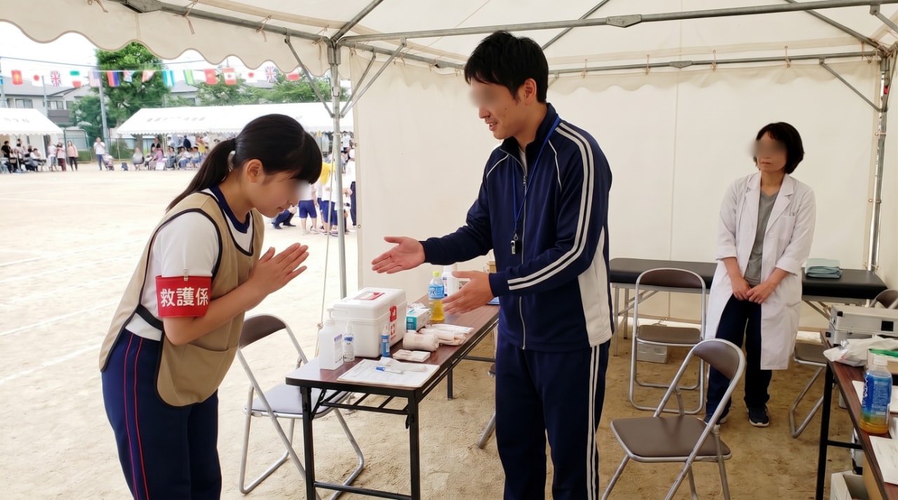 救護テント内で、救護係の腕章をつけた女性が深々と頭を下げて謝罪し、笑顔で手を広げて応じる男性教員とそれを見守る白衣の養護教諭。