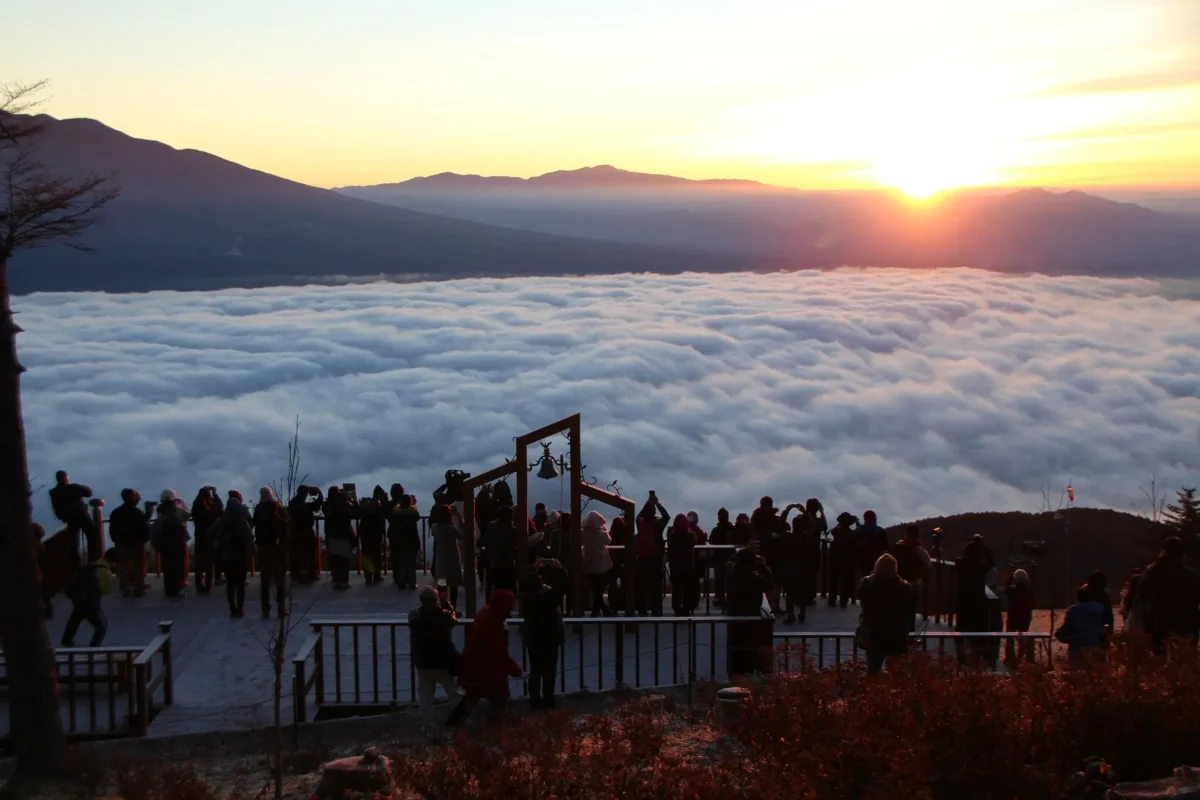 雲海の上に昇る朝日を眺める人々の写真。人々は展望台のような場所に集まり、カメラやスマートフォンでその光景を撮影している。遠くには山々がシルエットとして見え、空はオレンジ色に染まっている。