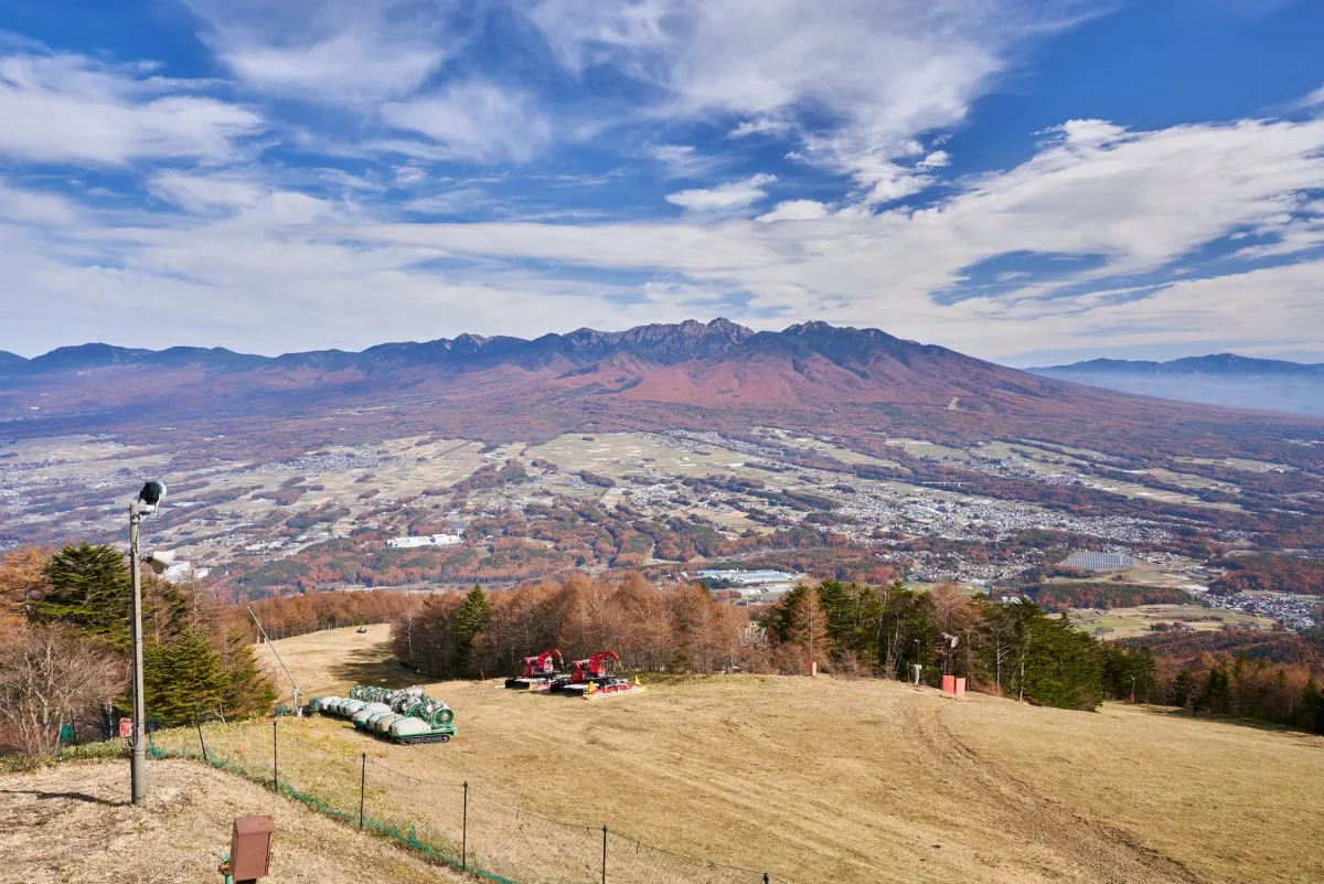 山の頂上から撮影された風景写真。遠くには山々が連なり、その麓には街並みが広がっている。手前には草木が生い茂り、雪山整備用の車両が配置されている。青い空には雲が広がり、自然の壮大さを感じさせる。