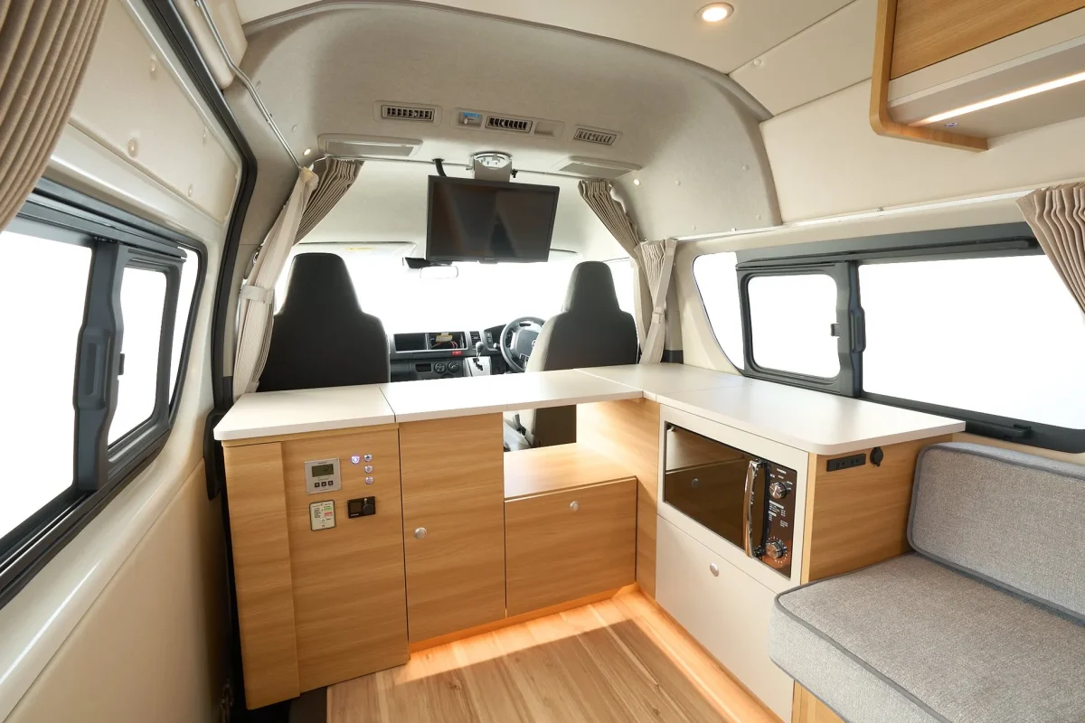 車内インテリアの写真です。木目調の家具が配置され、キッチンカウンター、テレビ、窓、カーテン、座席などが写っています。洗練されたデザインで、車中泊や移動空間としての快適さを感じさせます。