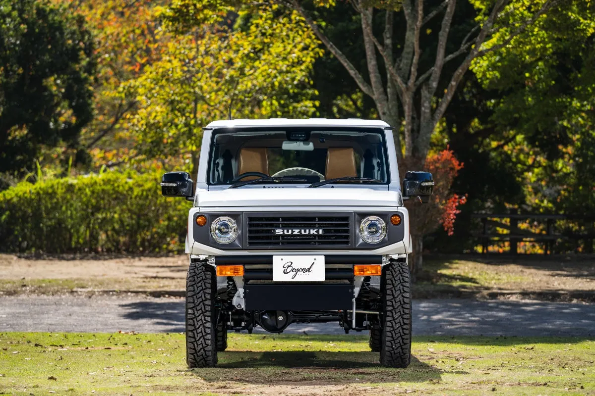 白いスズキジムニーの正面写真。背景には木々があり、車は草の上に駐車されている。