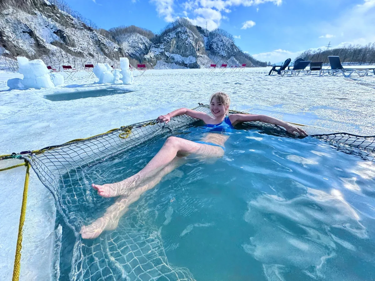 女性が水着を着て、雪景色の中で水風呂に入っている様子。周りには雪や氷があり、背景には山々が見える。