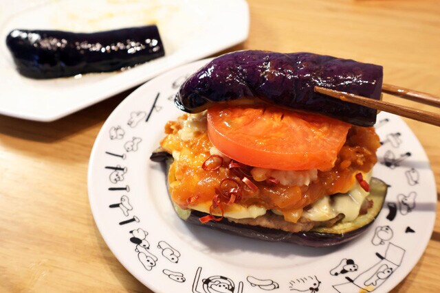 (4)仕上げ! 焼きナスの上にモスチーズバーガーの中身をのせ、その上にも焼きナスをのせれば完成! ただし、そのままかぶりつくと確実に汁が垂れまくる。再び包み紙に戻して食べるとよい