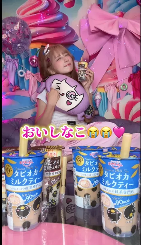 女性がタピオカミルクティーを持ち、クッションを抱きしめている写真。背景はピンクと紫を基調とした装飾が施されている。