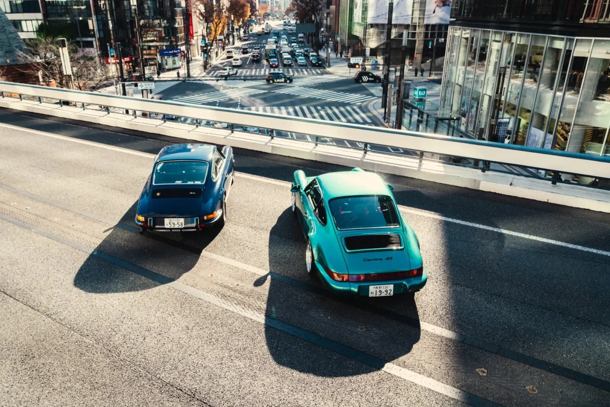 高架道路を走行する2台のヴィンテージポルシェ(青とティールグリーン)を上空から捉えた写真です。下には多くの車が行き交う賑やかな都市の交差点が広がっており、建物や横断歩道が見えます。日差しが強く、車や欄干の影が長く伸びています。