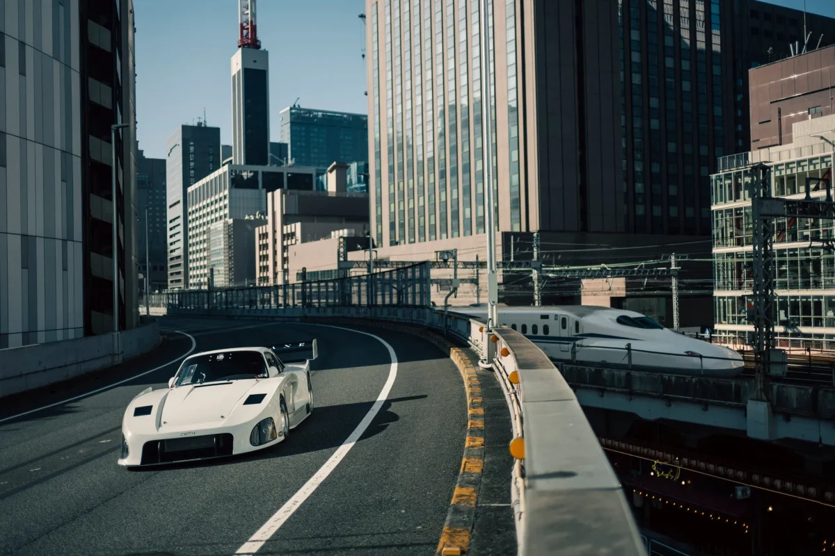 白いスポーツカーが都市の高架道路を走行し、その背景には新幹線と高層ビルが立ち並ぶ、現代的な日本の都市風景を捉えた一枚です。